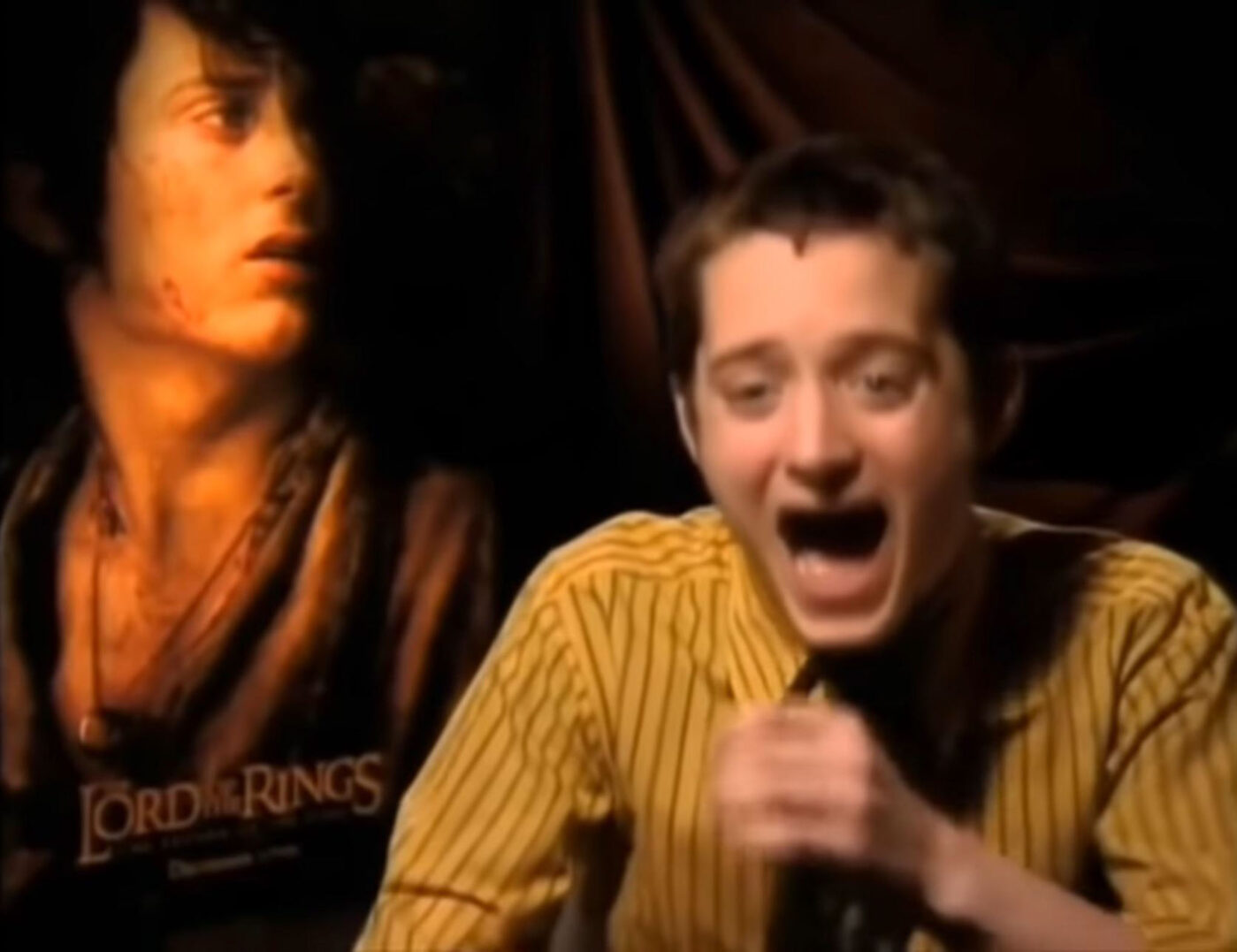 Elijha Wood laughing while being interviewed, with lord of the rings poster behind him.